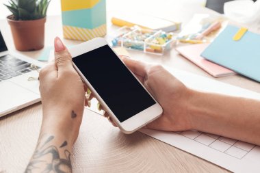 dizüstü bilgisayar ve kırtasiye ile ahşap masa arkasında oturan kadın kırpılmış görünümü, elinde akıllı telefon tutarak