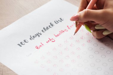 cropped view of woman writing notes on 100 days check list, holding green pen in hand clipart