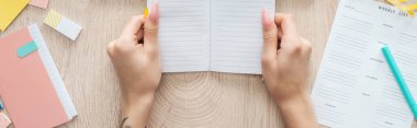 cropped view of woman holding notepad in hands over wooden table with weekly list and stationery clipart