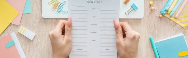 cropped view of woman holding weekly list over wooden table with laptop and stationery clipart