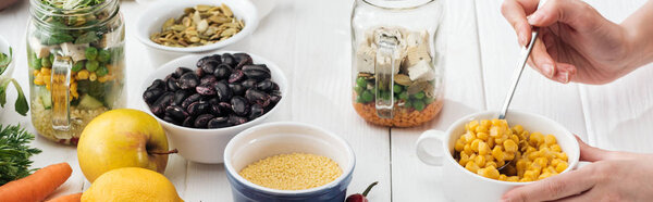 cropped view of woman adding corn in glass jar with salad on wooden white table, panoramic shot
