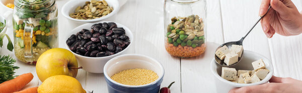 cropped view of woman adding tofu cheese in glass jar on wooden white table, panoramic shot