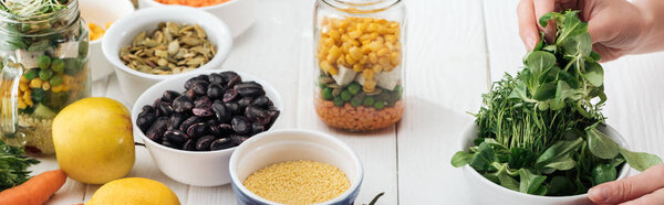 cropped view of woman taking greens from plate on wooden white table, panoramic shot