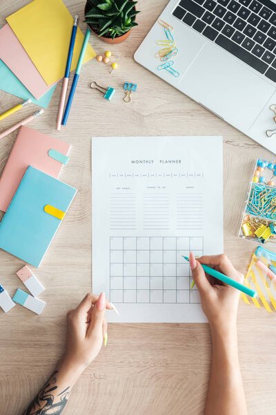 cropped view of woman sitting behind wooden table with laptop and stationery, writing notes in monthly planner