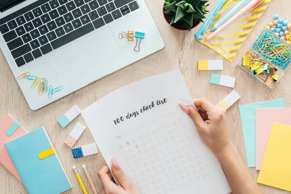 cropped view of woman sitting behind wooden table with laptop and stationery, holding in hands 100 days check list