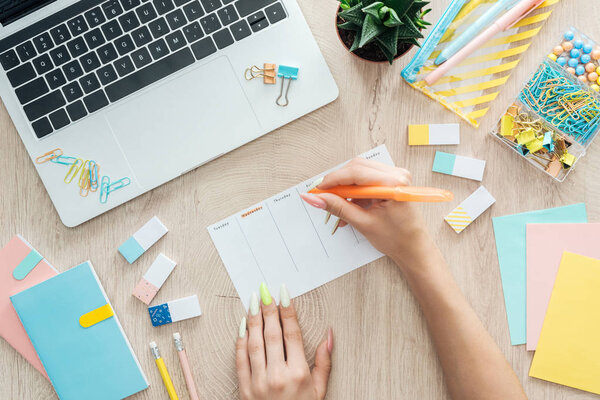cropped view of woman holding marker in hand, writing notes in planner, sitting behind wooden table with laptop, stationery and plant