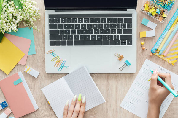 обрезанный вид женщины, сидящей за деревянным столом с ноутбуком, канцелярскими принадлежностями и еженедельным списком
