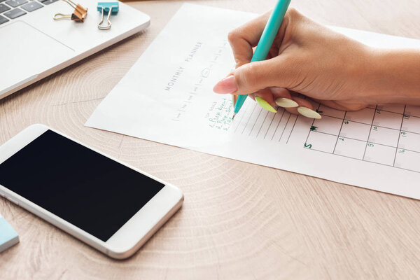 cropped view of woman writing notes in planner, sitting behind wooden table with laptop and smartphone