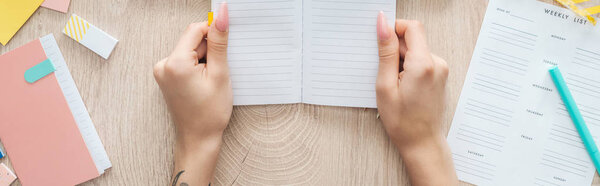 cropped view of woman holding notepad in hands over wooden table with weekly list and stationery