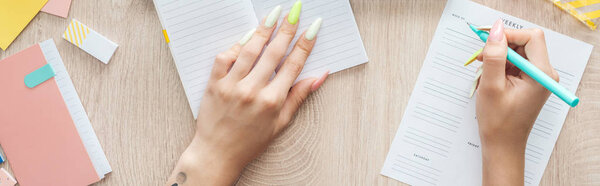 cropped view of woman writing in weekly list, sitting behind wooden table with notepad and stationery