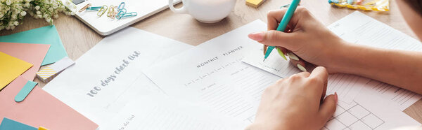 cropped view of woman holding pen in hand, writing notes in planners, sitting behind wooden table with flowers and stationery