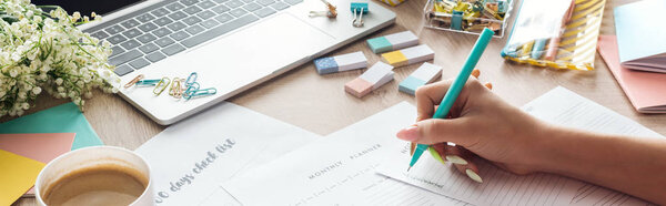 cropped view of woman holding pen in hand, writing notes in planners, sitting behind wooden table with flowers and stationery