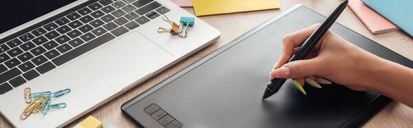 обрезанный вид женщины, работающей с графическим планшетом, сидящей за деревянным столом с ноутбуком и канцелярскими принадлежностями
