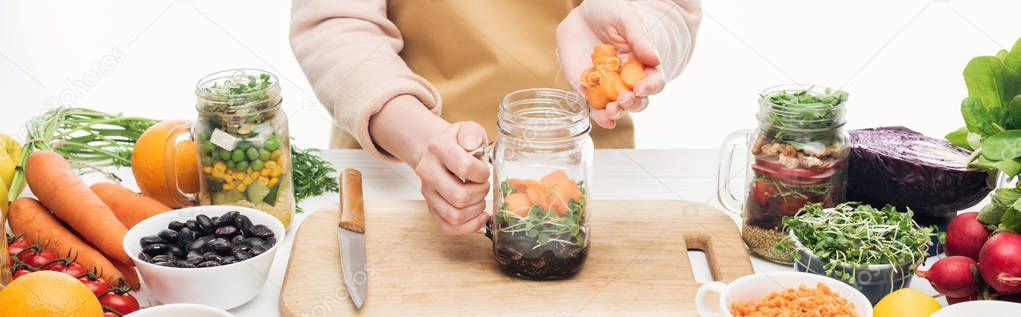 cropped view of woman in apron adding carrots to glass jar with beans on wooden table isolated on white, panoramic shot