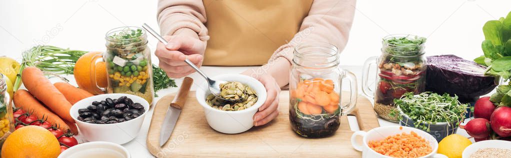 cropped view of woman in apron adding pumpkin seeds from bowl in glass jar on wooden table isolated on white, panoramic shot