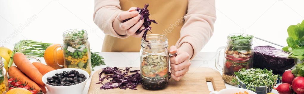 cropped view of woman in apron adding red cabbage in glass jar on wooden table isolated on white, panoramic shot