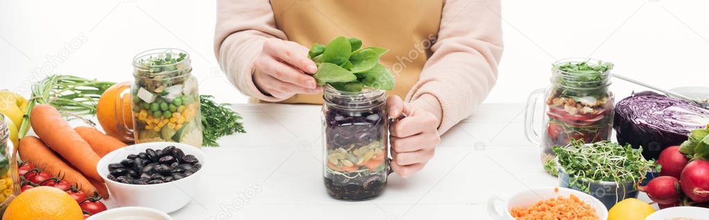 partial view of woman in apron holding glass jar with salad and green leaves on wooden table isolated on white, panoramic shot