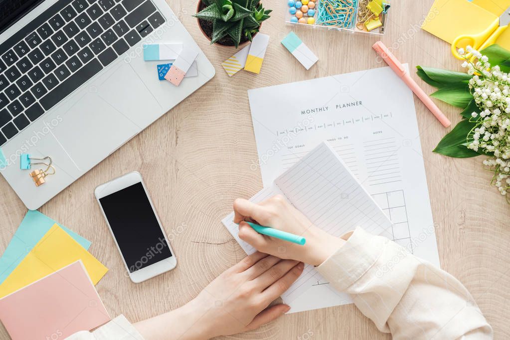 cropped view of woman sitting behind wooden table with smartphone, laptop and stationery, writing in notepad