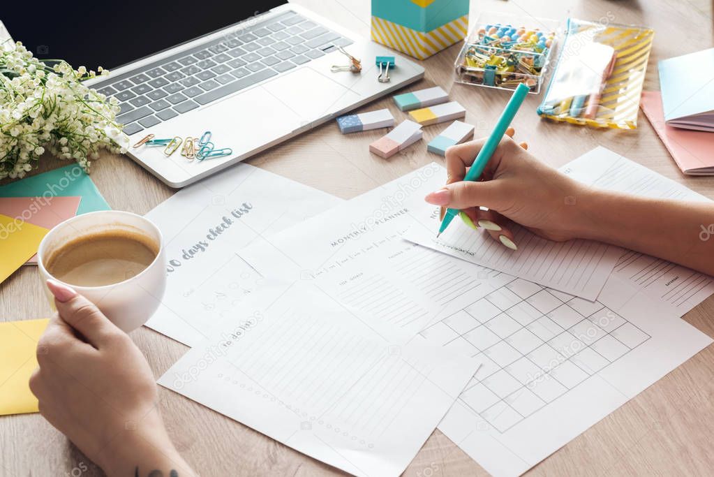 cropped view of woman holding cup of coffee in hand, sitting behind wooden table with laptop and stationery, writing in paper planners