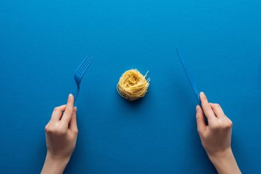 mavi arka plan üzerinde orta erişte makarna ile mavi plastik çatal ve bıçak tutan kadının kırpılmış görünümü