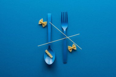 top view of spaghetti on blue plastic spoon upside down and fork near two kinds of pasta on blue background clipart