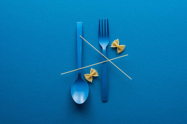 top view of spaghetti on blue plastic spoon upside down and fork near farfalle pasta on blue background