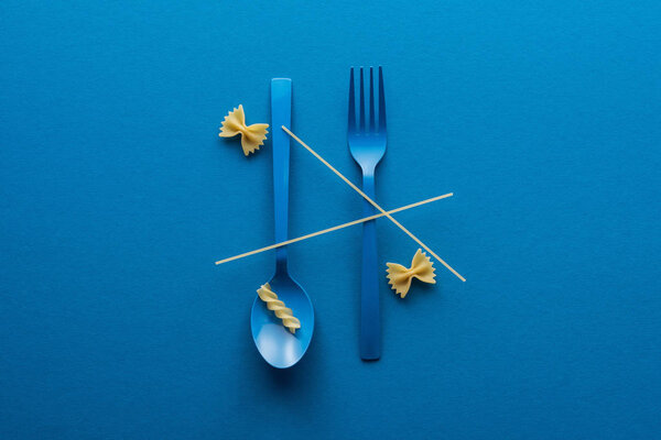 top view of spaghetti on blue plastic spoon upside down and fork near two kinds of pasta on blue background