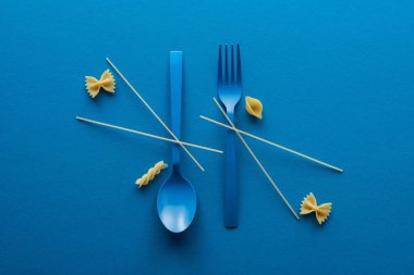 flat lay of spaghetti on blue plastic spoon upside down and fork near different kinds of uncooked pasta on blue background clipart
