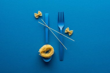 vermicelli pasta on blue plastic spoon with spaghetti on fork and spoon near farfalle pasta on blue background clipart