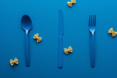 mavi arka plan etrafında pişmemiş farfalle makarna ile mavi plastik kaşık, bıçak ve çatal