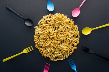 siyah arka plan üzerinde renkli çatal bıçak takımı ile çevrili pişmemiş makarna üst görünümü