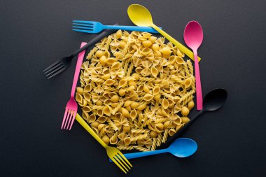 siyah arka plan üzerinde renkli kaşık ve çatal ile çevrili pişmemiş makarna üst görünümü