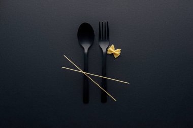 top view of spaghetti noodles on plastic cutlery near farfalle pasta on black background clipart