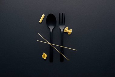 black plastic spoon, fork and four different kinds of pasta on black background clipart