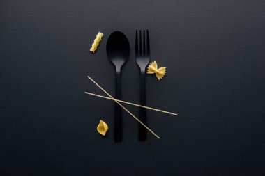 black plastic spoon, fork and four different kinds of pasta on black background clipart