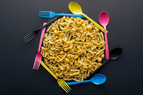 在黑色背景上用五颜六色的勺子和叉子包围未煮熟的意大利面的顶视图 — 图库照片