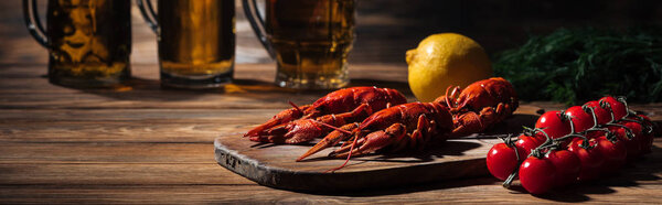 panoramic shot of red lobsters, tomatoes, dill, lemon and glasses with beer on wooden surface