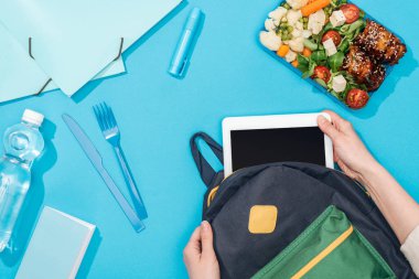 öğle yemeği kutusu, kırtasiye ve su şişesi yakınında sırt çantasında dijital tablet ambalaj kadın kırpılmış görünümü
