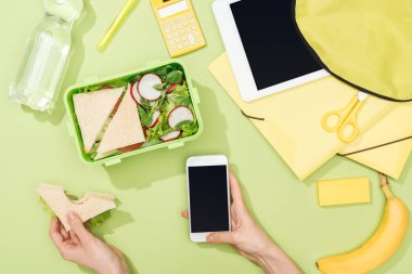 yemek, sırt çantası, dijital tablet, su şişesi ve kırtasiye ile öğle yemeği kutusu yakınında sandviç ve akıllı telefon ile kadın ellerin kırpılmış görünümü
