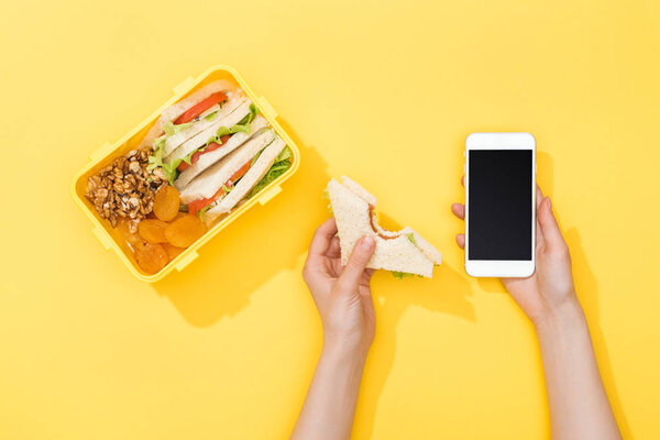 обрезанный вид женщины, держащей сэндвич и смартфон возле коробки с едой
