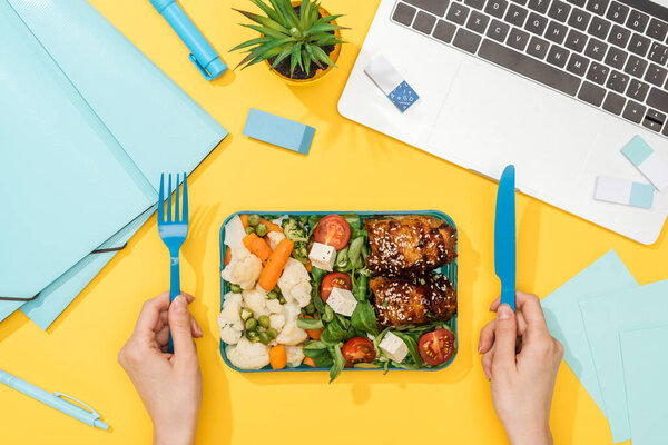 cropped view of woman holding fork over lunch box with food near laptop and office supplies
