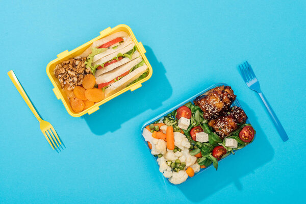 top view of plastic forks near lunch boxes with food on blue background