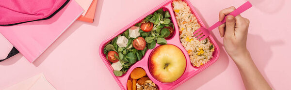 panoramic shot of woman holding fork over lunch box near backpack and stationery