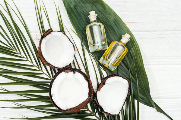 top view of coconut oil in bottles on green palm leaves on white wooden surface