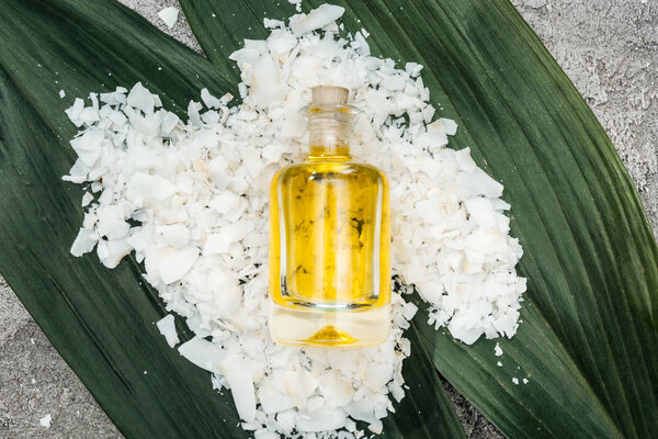 top view of coconut oil in bottle on coconut shaving and palm leaves on grey textured background