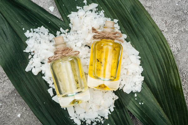 top view of coconut oil in bottles on coconut shaving and palm leaves on grey textured background