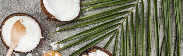 top view of coconut shavings with wooden spoon on grey textured background with palm leaf and coconuts, panoramic shot