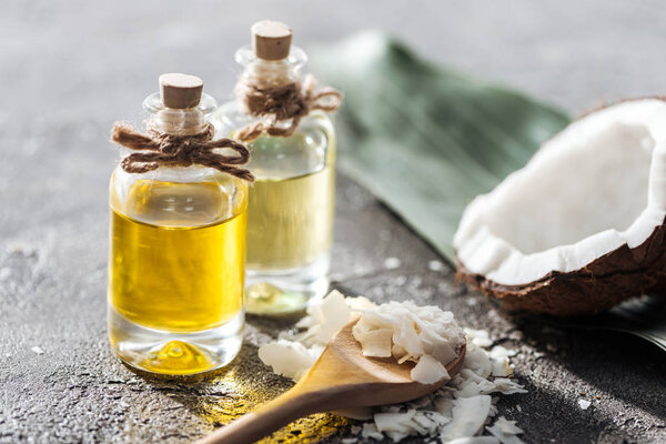 selective focus of bottles with coconut oil near coconut half and shavings in wooden spoon on grey background