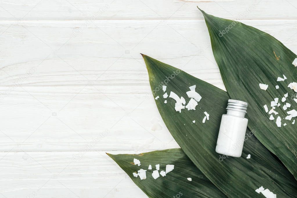 top view of coconut beauty product in bottle on green palm leaves with coconut flakes on white wooden surface