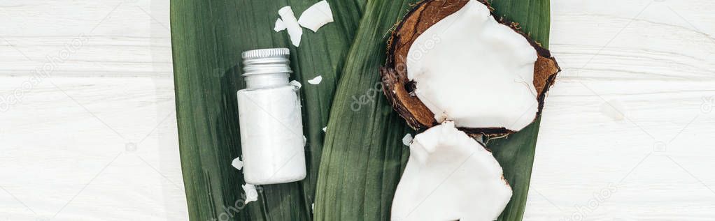 top view of coconut beauty product in bottle on green palm leaves with coconut flakes on wooden surface, panoramic shot